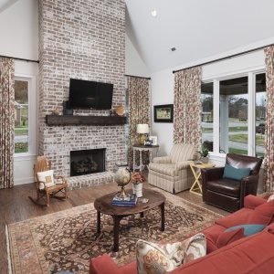 A cozy living room with a brick fireplace, mounted TV, and patterned curtains. There's a red sofa, armchairs, and a wooden rocking chair. A rug and coffee table complete the space.