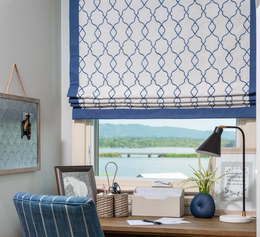 A home office with a wooden desk showcases a blue patterned chair, a lamp, and decorative items. The window, adorned with a blue and white Roman shade—a perfect example of why window treatments matter—offers a stunning view of the lake and hills.