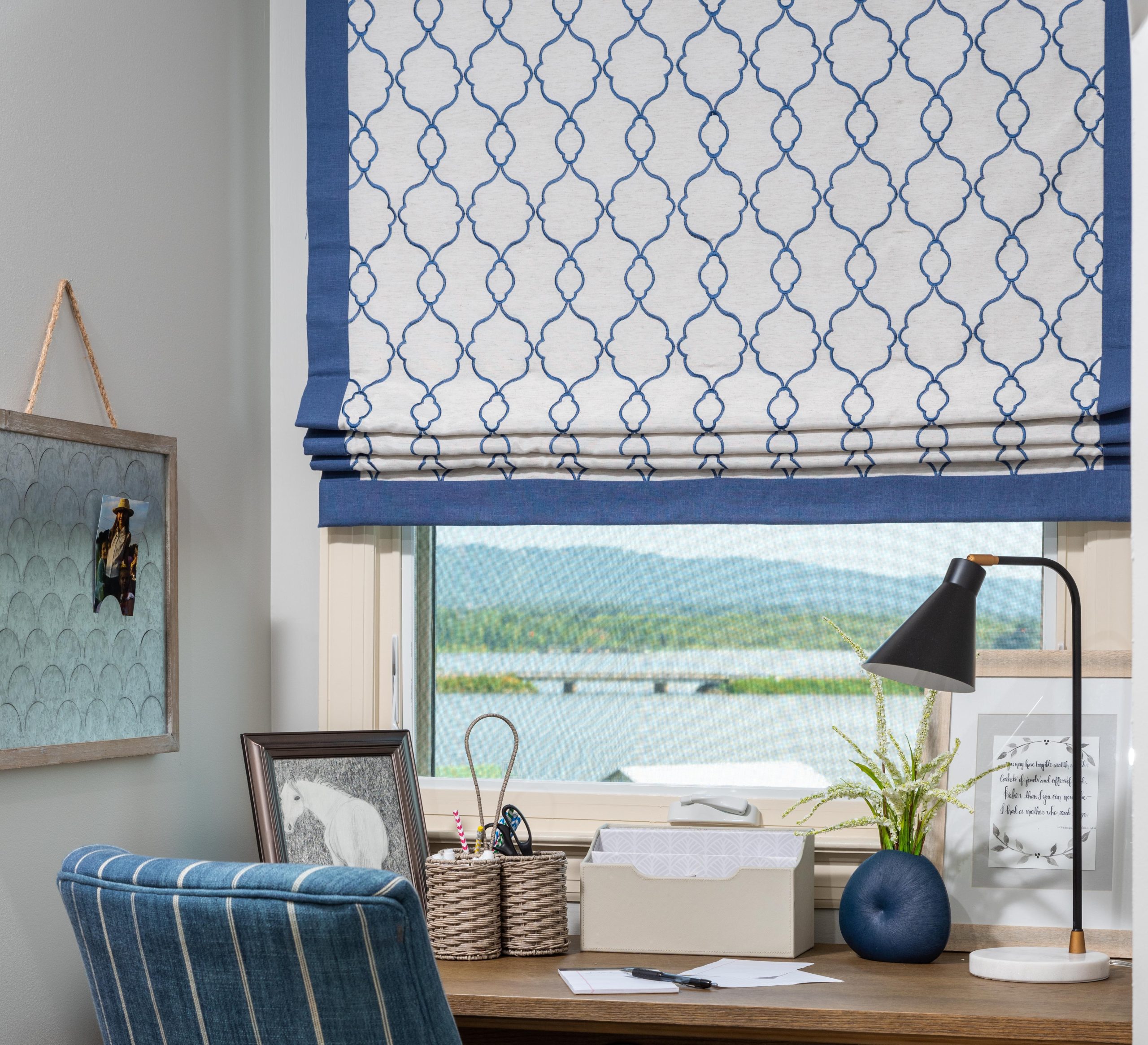 A home office with a wooden desk showcases a blue patterned chair, a lamp, and decorative items. The window, adorned with a blue and white Roman shade—a perfect example of why window treatments matter—offers a stunning view of the lake and hills.