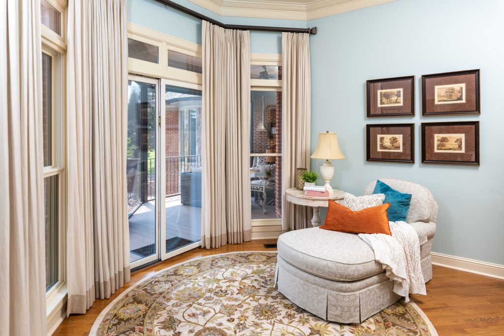 Cozy sitting area with a patterned round rug, a plush armchair adorned with orange and blue cushions, and a side table with a lamp. Curtains frame large windows overlooking the patio, transforming your home into your haven.