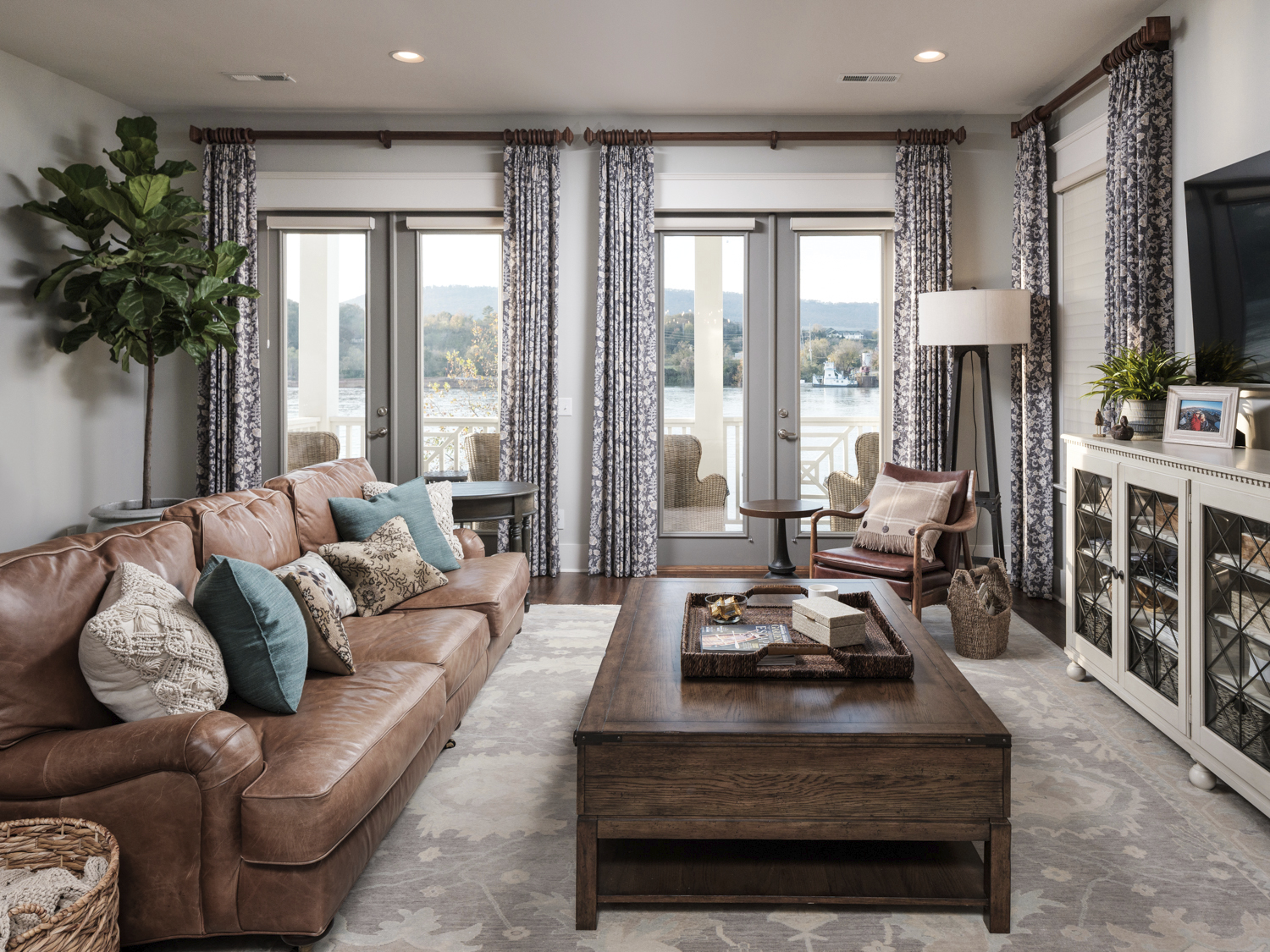 A cozy living room with a brown leather sofa, various pillows, a wooden coffee table, a large potted plant, and a view of a lake through the windows with patterned curtains.