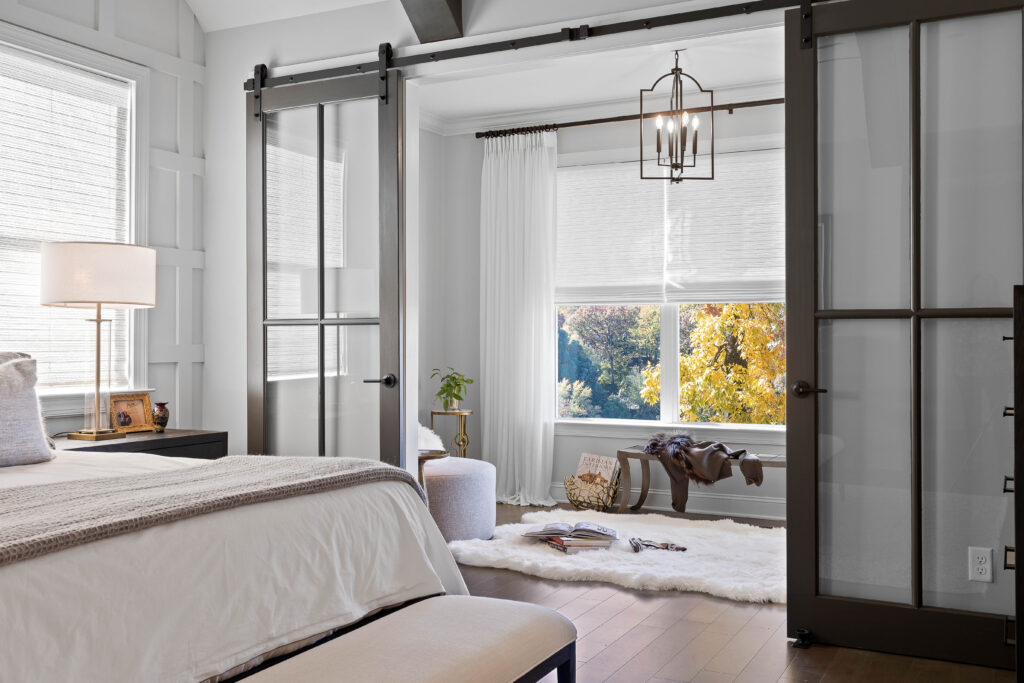 A modern bedroom with glass sliding doors opens to a bright sitting area with large windows, a white rug, a bench, and a contemporary chandelier.