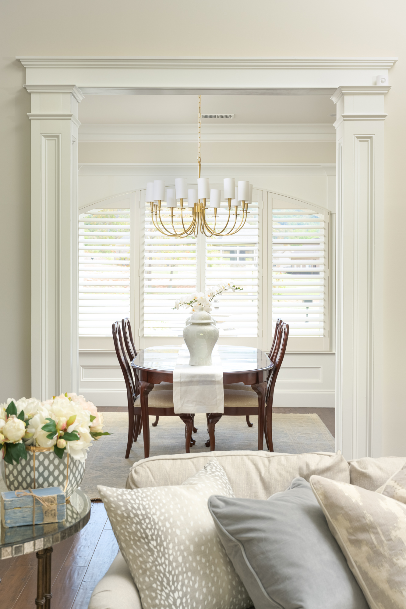 A dining room with a dark wood table and chairs, white vase centerpiece, and gold chandelier, viewed through an archway from a living area with neutral-toned pillows and decor.