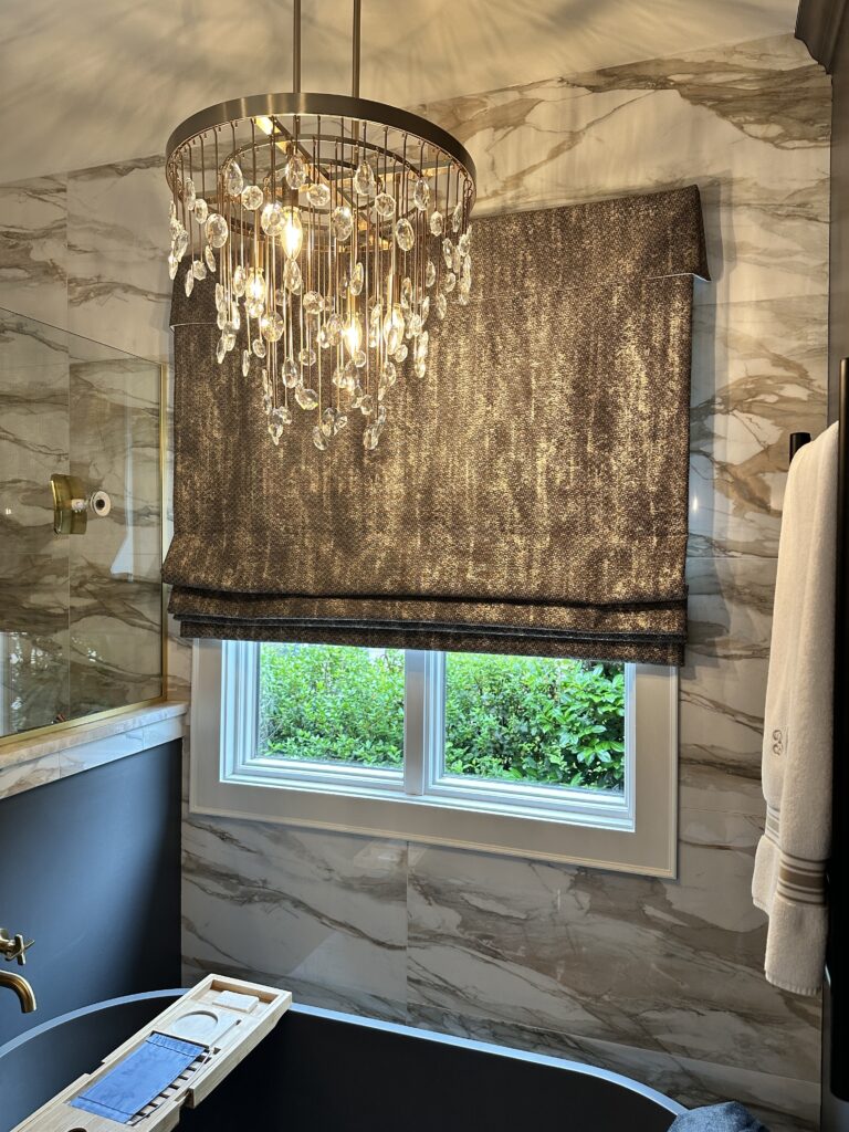 Modern bathroom with marble walls, a crystal chandelier, a window with a textured Roman shade, and a black tub with a bath tray beneath the window.