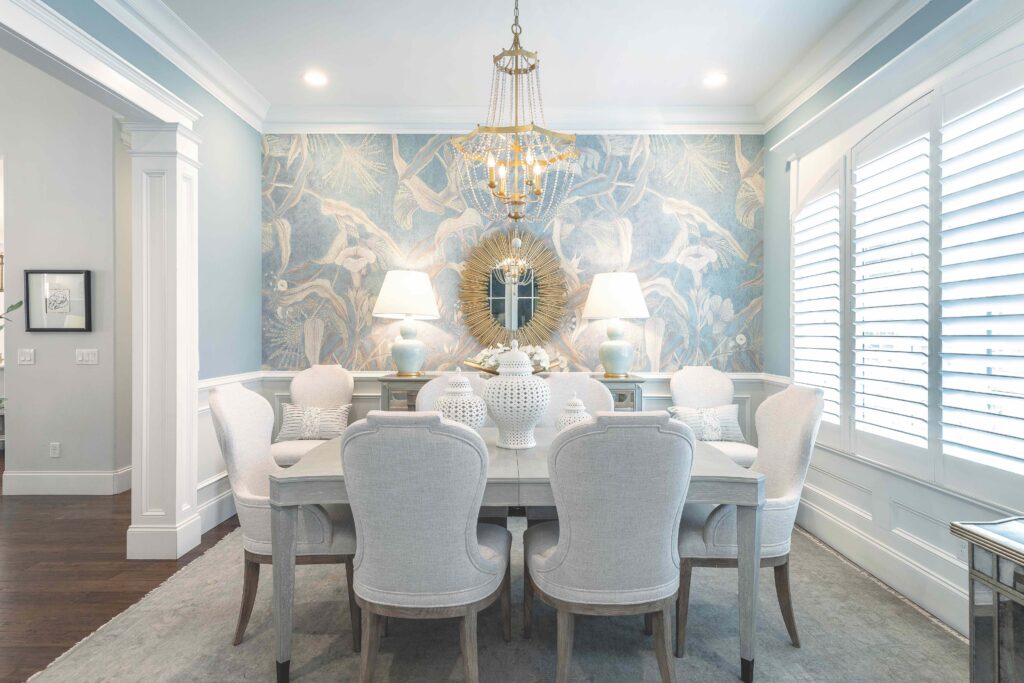 Elegant dining room with a rectangular table, eight upholstered chairs, two lamps, decorative vases, a gold sunburst mirror, and a chandelier, set against a blue patterned accent wall.